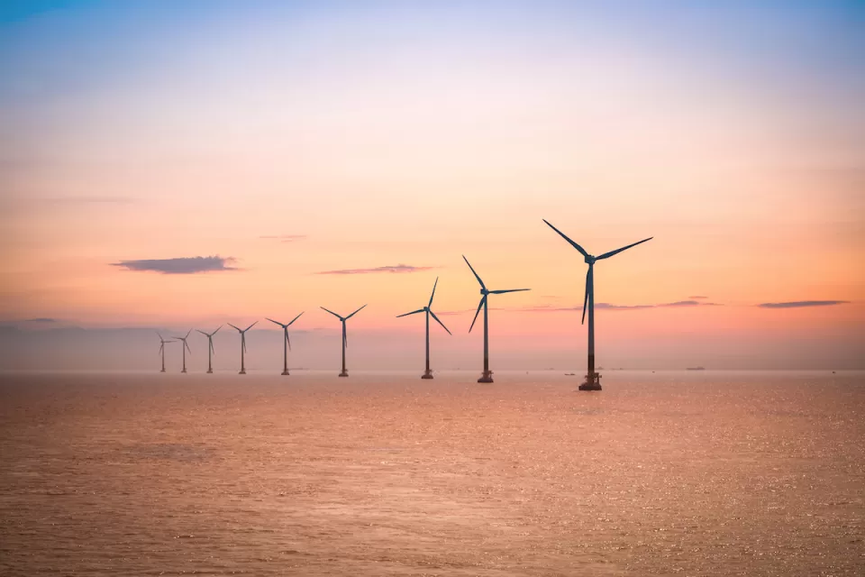 Row of offshore wind turbines in large water body against sunrise or sunset sky.
